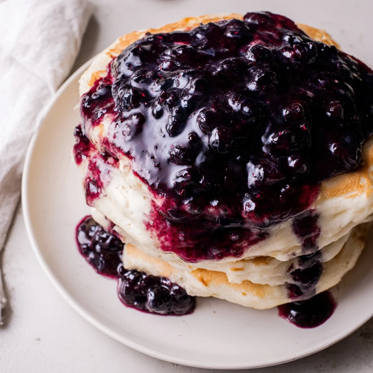 Fluffy Lemon Ricotta Pancakes with Blueberry Syrup are stacked high, drizzled with vibrant, glossy syrup, and dusted with powdered sugar on a rustic breakfast table.